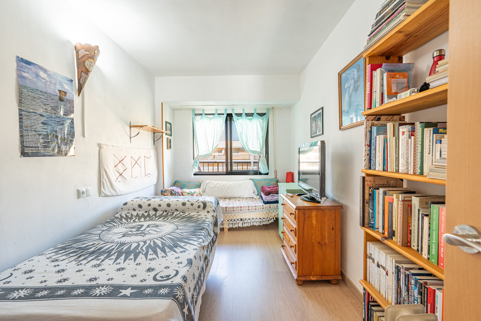 Chambre avec lit simple, étagères en bois et commode. Comprend un sol stratifié et une fenêtre avec des rideaux bleu clair offrant une lumière naturelle.