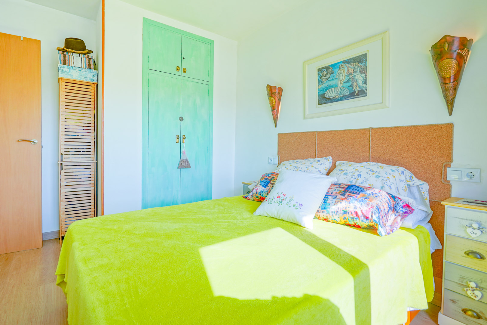 Chambre avec tête de lit en liège, armoire encastrée vert menthe et œuvre d'art. La lumière naturelle éclaire le couvre-lit citron vert et la table de chevet.