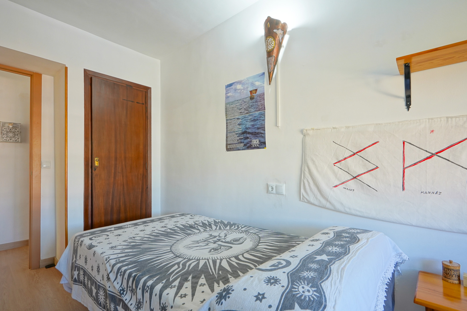 Chambre équipée d'un lit simple avec couvre-lit soleil, murs blancs avec une applique en cuivre travaillée et une porte en bois massif sombre.