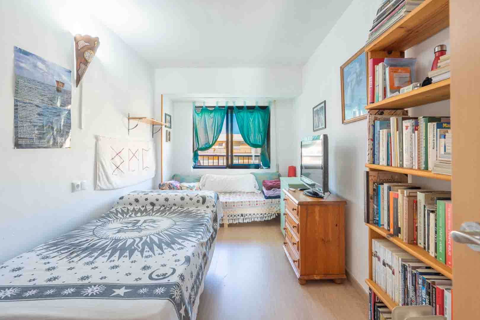 Chambre lumineuse avec lit simple, bibliothèque en bois et commode avec télévision. La lumière naturelle pénètre par une fenêtre ornée de rideaux turquoise.