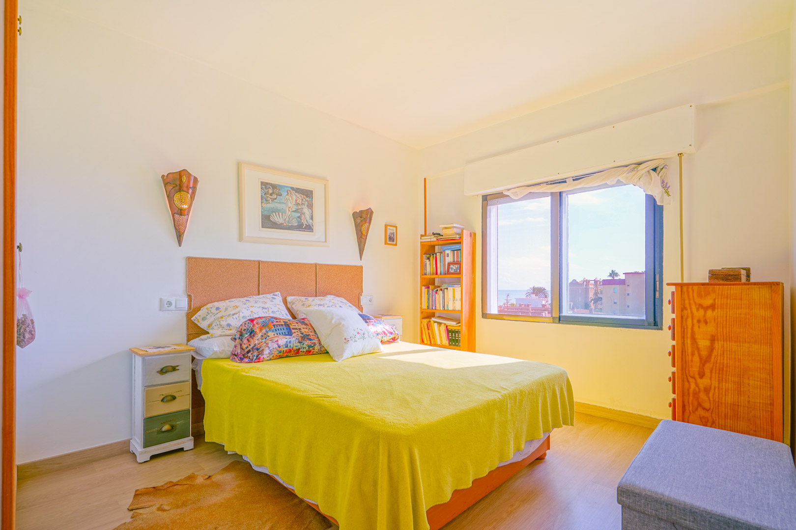 Chambre lumineuse avec tête de lit en liège, couvre-lit jaune et meubles en bois. Une grande fenêtre offre une lumière naturelle et une vue dégagée.