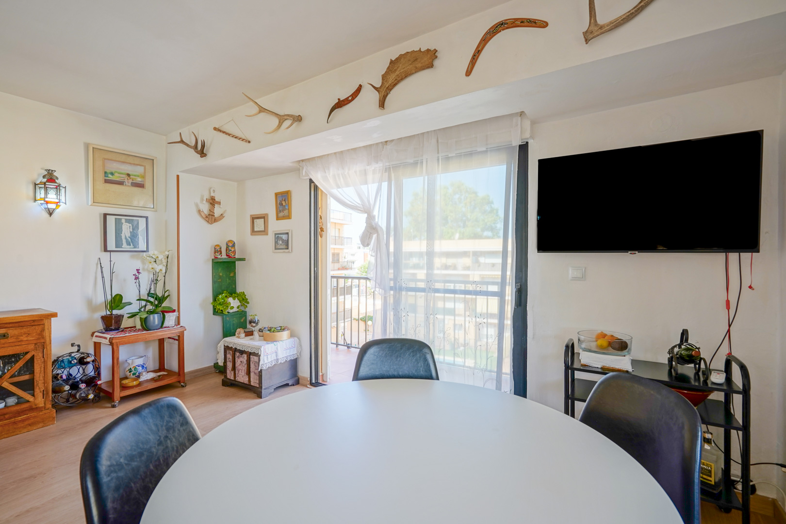 Coin repas avec table circulaire blanche, télévision murale et accès au balcon. La décoration originale inclut des bois de cerf et des boomerangs fixés au mur.