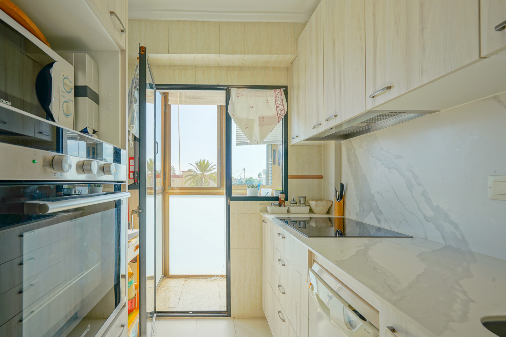 Cuisine équipée avec placards en bois clair, plan de travail effet marbre et accès direct à un balcon offrant une vue sur des palmiers.