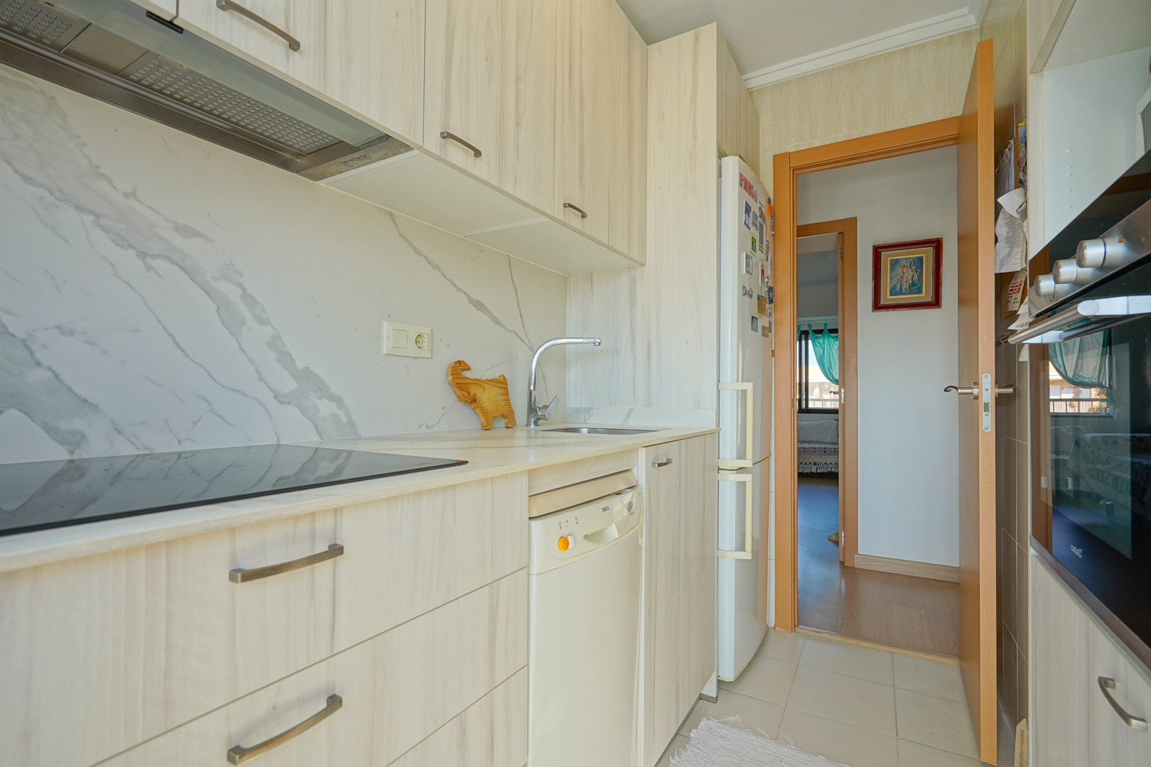 Cuisine fonctionnelle avec meubles en bois clair, crédence effet marbre, plaque à induction et électroménager intégré ouvrant sur le couloir.