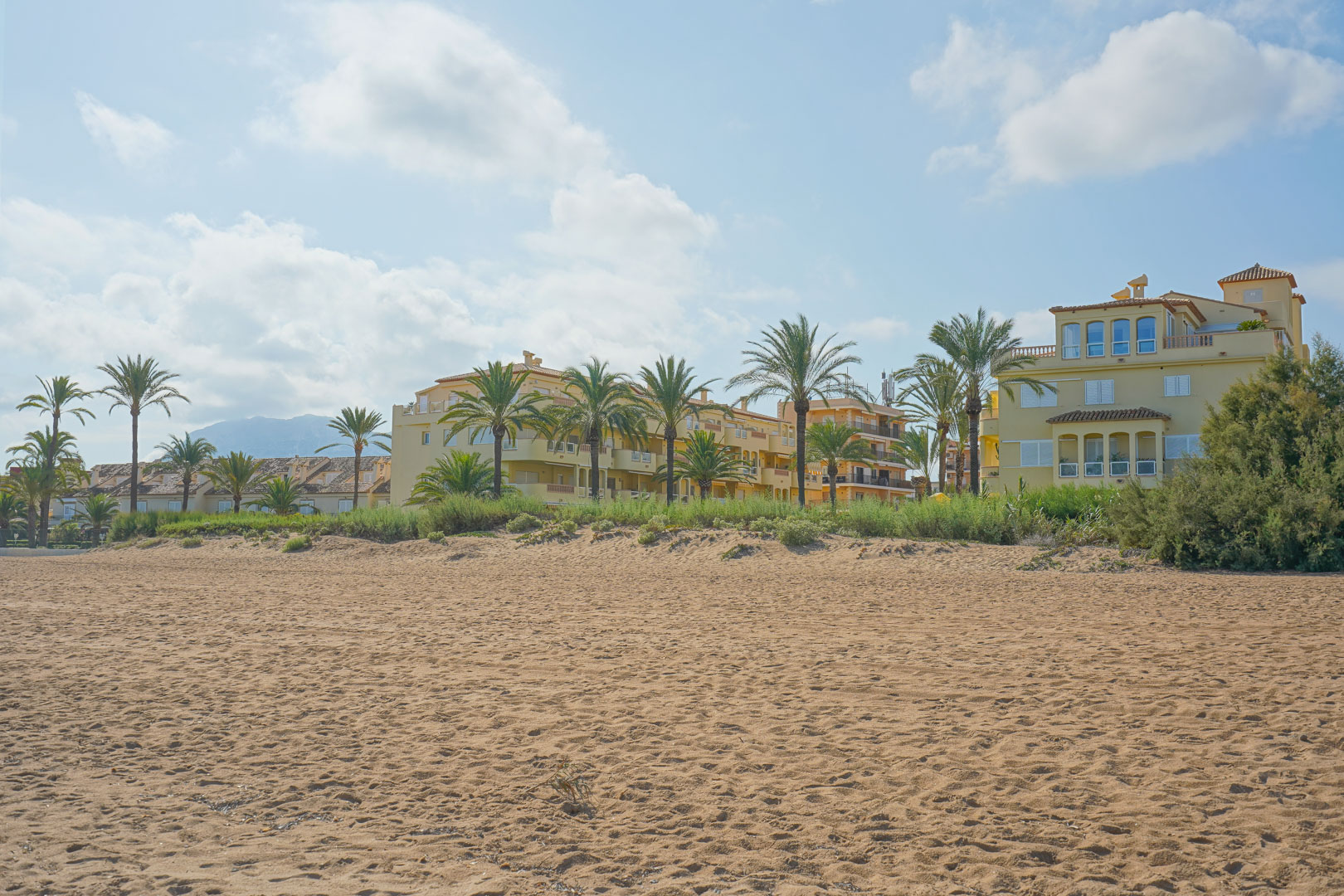 Large plage de sable bordant des résidences de style méditerranéen avec palmiers et vue sur les montagnes au loin sous un ciel clair.