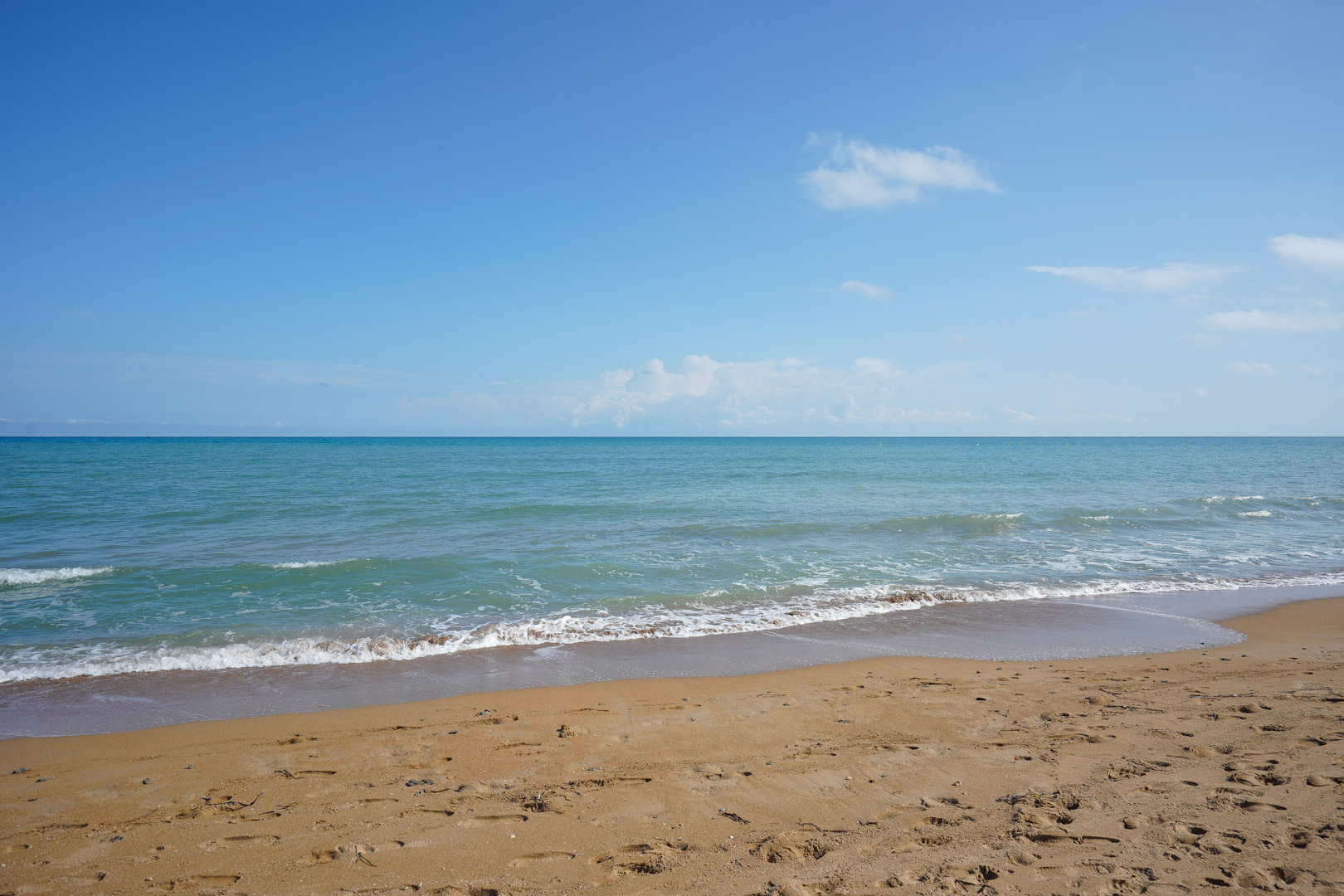 Large plage de sable doré bordée par des vagues turquoise sous un ciel bleu clair. Le rivage offre une vue paisible sur les eaux calmes de la Méditerranée.