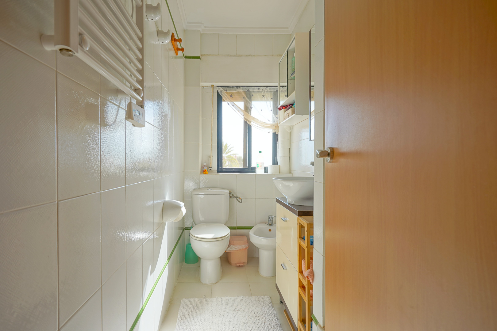 Salle de bain fonctionnelle avec carrelage blanc, bidet, toilettes et meuble vasque. Une fenêtre laisse entrer la lumière naturelle et offre une vue sur l'extérieur.