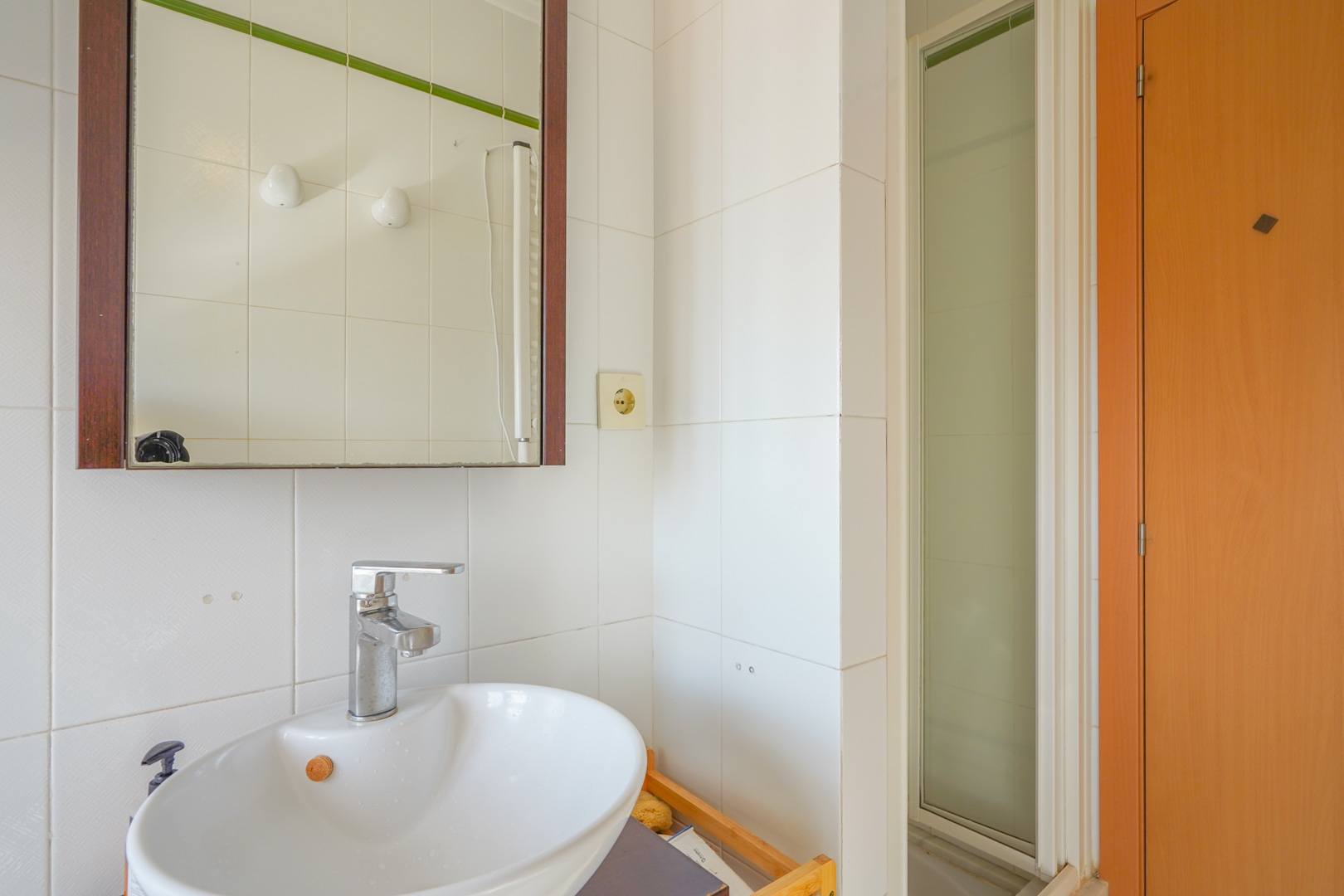 Salle de bain fonctionnelle avec vasque en céramique blanche, robinetterie chromée et miroir à cadre en bois, à côté d'une douche en verre dépoli.