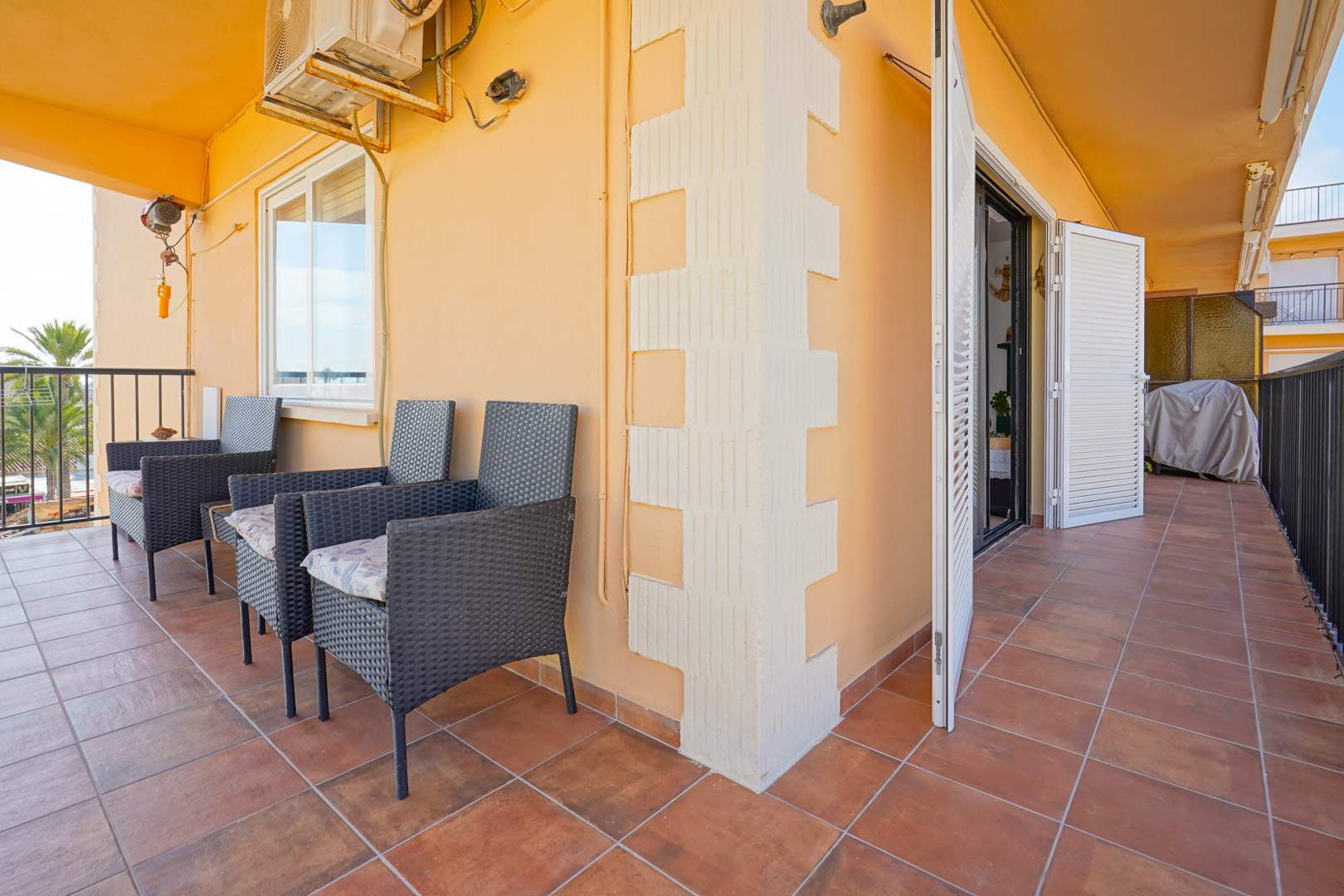 Terrasse couverte avec carrelage en terre cuite, fauteuils en résine tressée et volets blancs. L'espace est équipé d'une unité de climatisation murale.