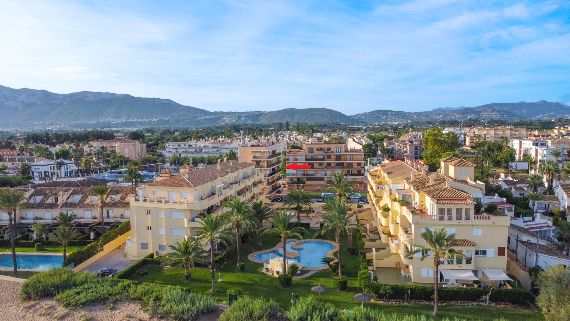 Vue aérienne du complexe avec piscine et jardins. L'emplacement précis de l'appartement est indiqué, offrant une perspective sur l'environnement montagneux.