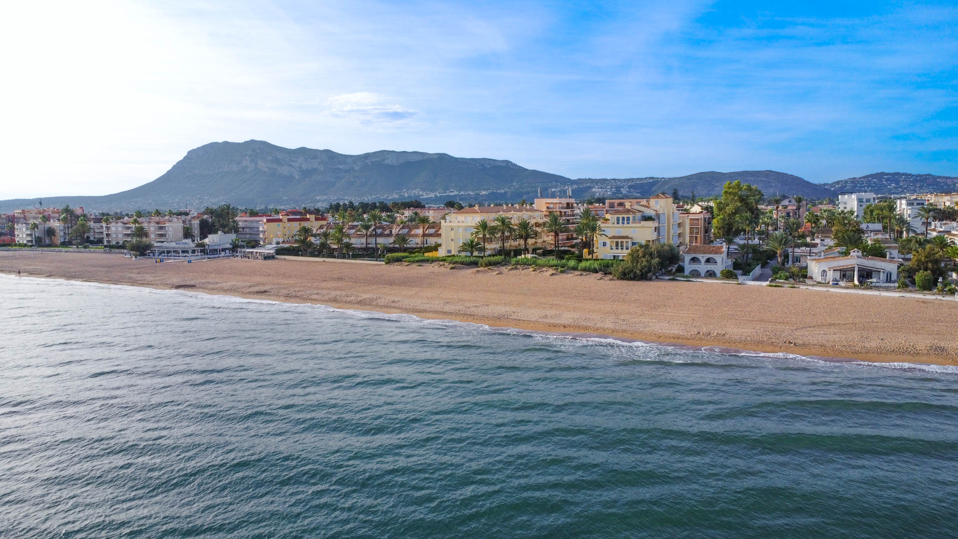 Vue aérienne d'une plage de sable bordée de complexes résidentiels et de palmiers, avec une chaîne de montagnes en arrière-plan sous un ciel dégagé.