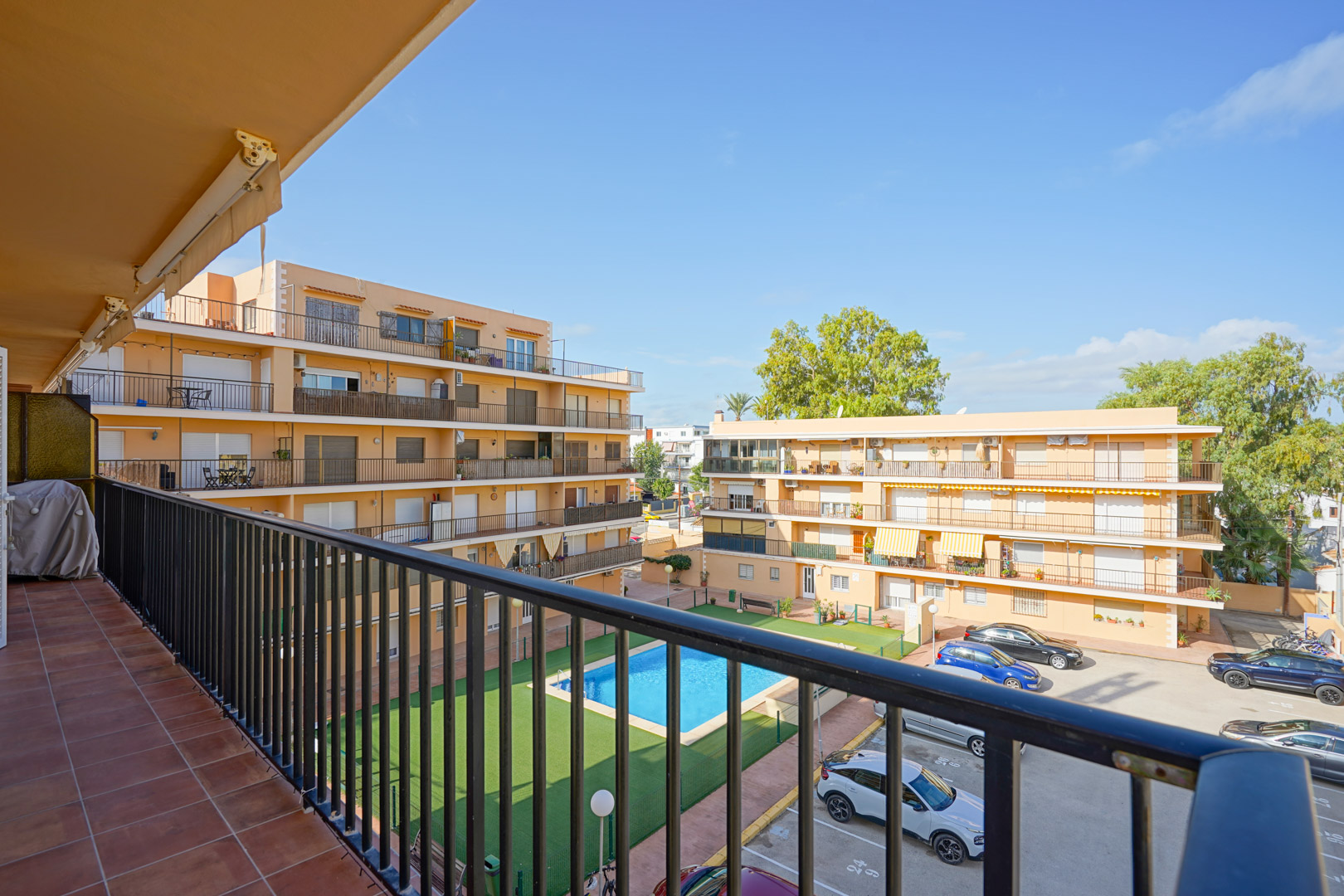 Vue depuis un balcon privé carrelé avec garde-corps métallique, donnant sur la piscine commune, la pelouse artificielle et le parking de la résidence.