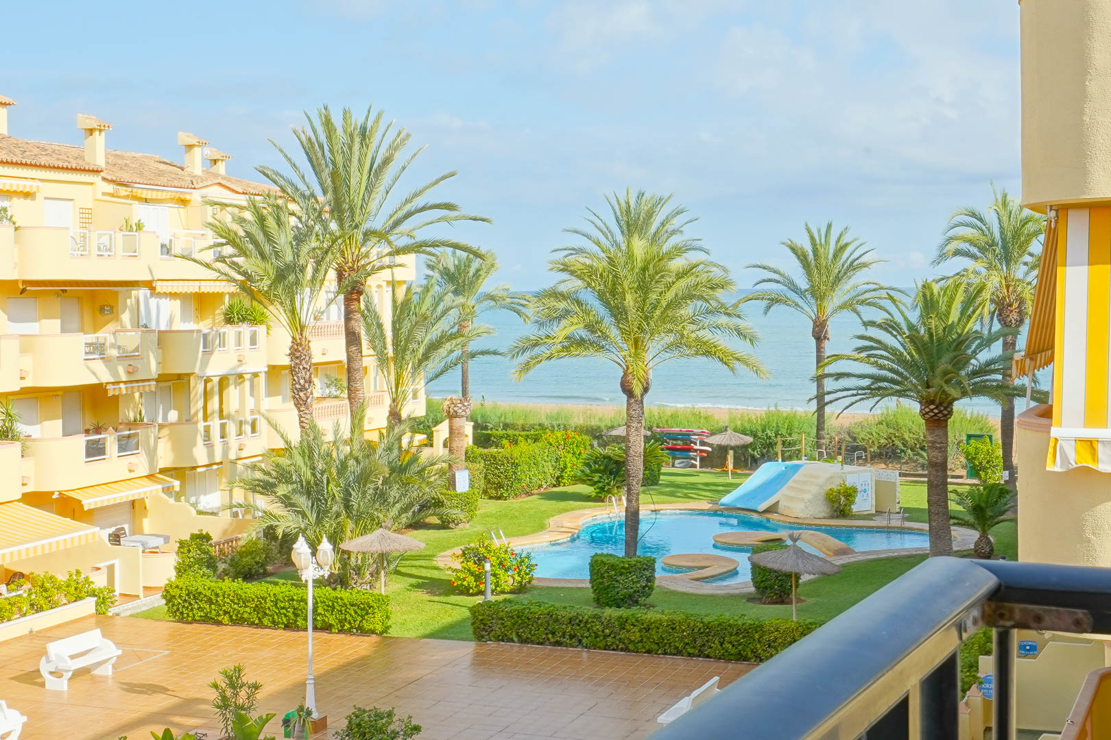 Vue depuis un balcon privé sur un jardin paysager avec palmiers, piscine avec toboggan et panorama dégagé sur la mer Méditerranée.