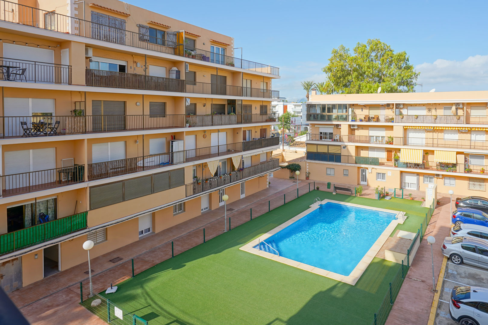 Vue du complexe résidentiel comprenant une piscine rectangulaire, une zone de gazon artificiel clôturée et des bâtiments avec balcons et parking extérieur.