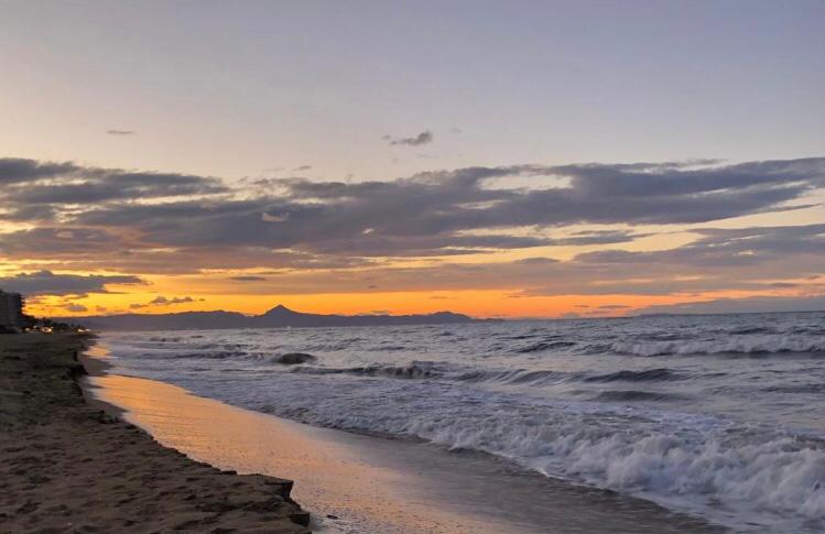 Vue du coucher de soleil sur une plage de sable avec des vagues et des silhouettes de montagnes sous un ciel nuageux orangé.