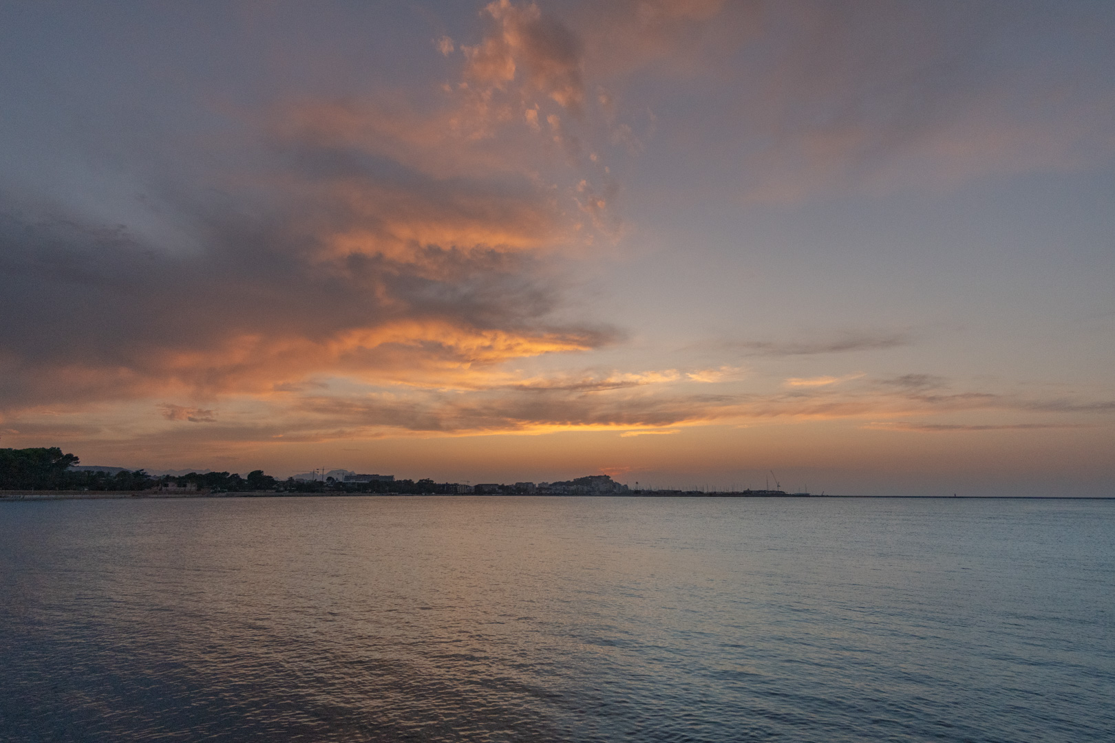 Vue panoramique de la côte méditerranéenne au coucher du soleil, montrant des nuages orangés et des eaux calmes reflétant le ciel crépusculaire sur la ville au loin.