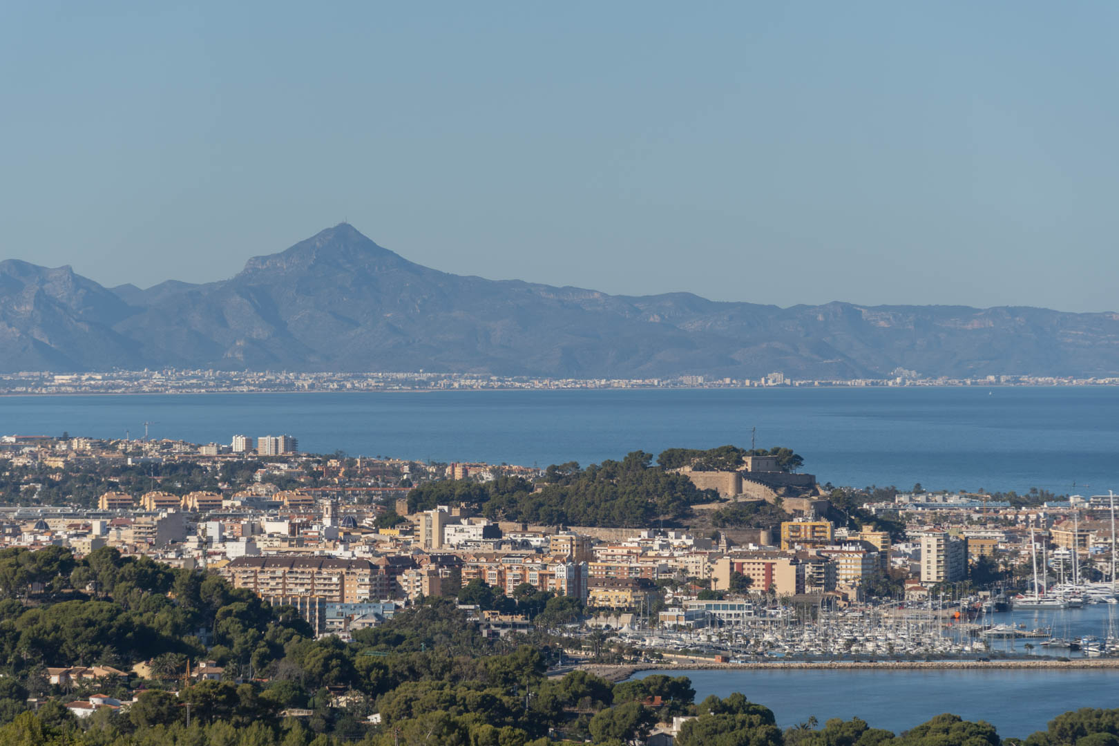 Vue panoramique de la ville côtière avec son château historique, le port de plaisance et une chaîne de montagnes en arrière-plan au-delà de la baie.