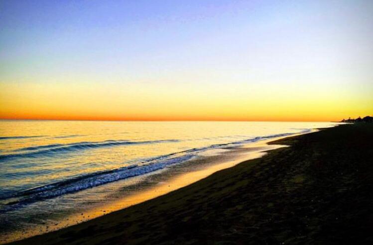 Vue panoramique d'une plage de sable au crépuscule, montrant des vagues légères et un dégradé de couleurs allant du bleu profond au jaune doré à l'horizon.