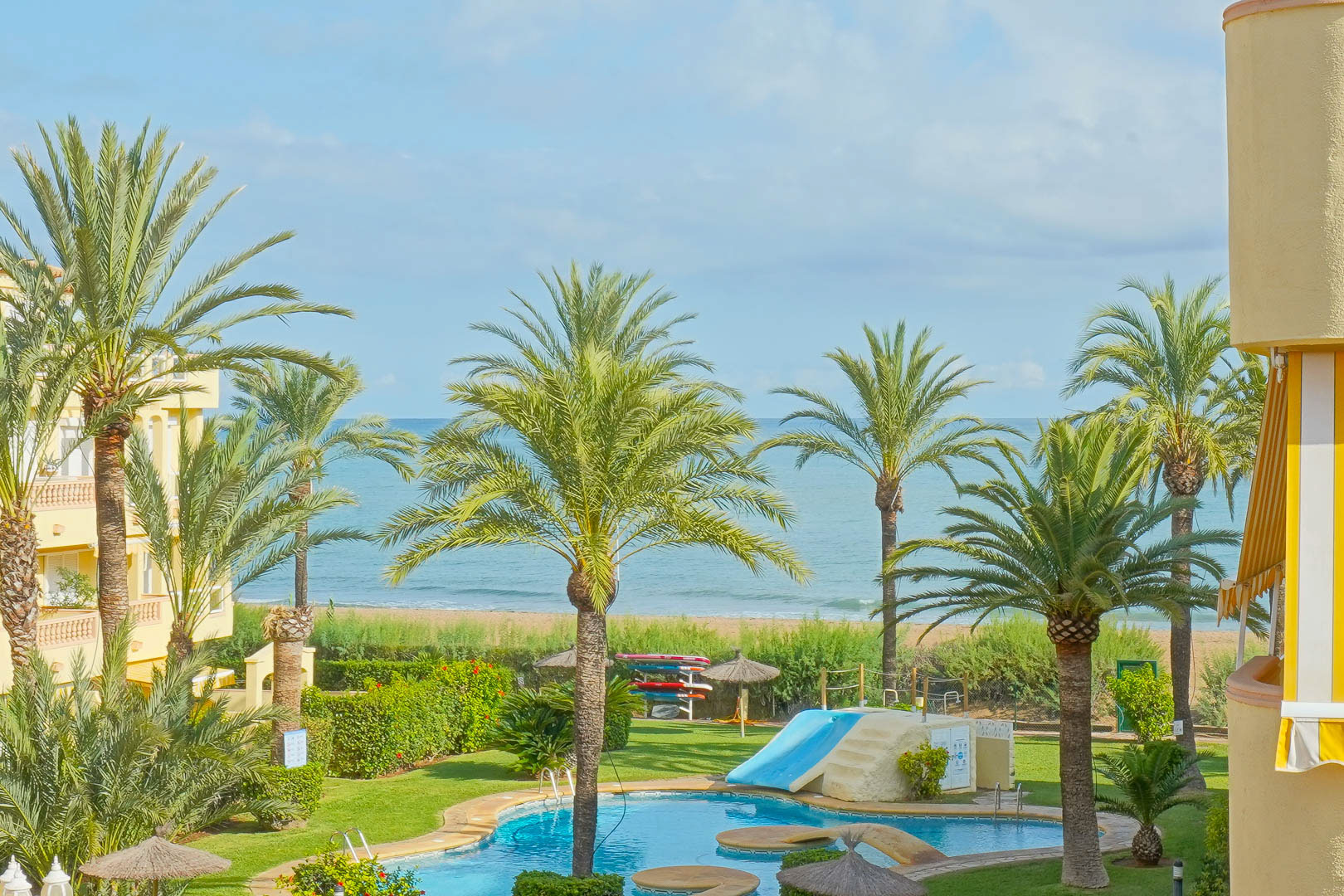 Vue sur la piscine commune avec toboggan, entourée de palmiers et de jardins, offrant un panorama direct sur la plage et la mer Méditerranée.
