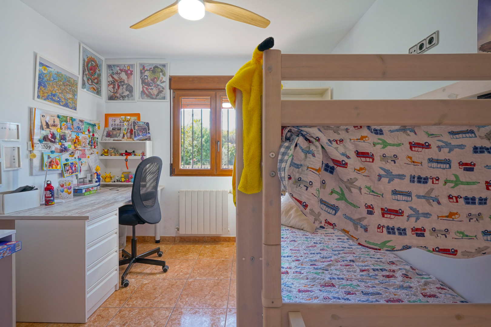 Chambre d'enfant avec lits superposés en bois, bureau blanc et fenêtre à cadre en bois. La pièce dispose d'un radiateur, d'un ventilateur de plafond et d'un sol carrelé.