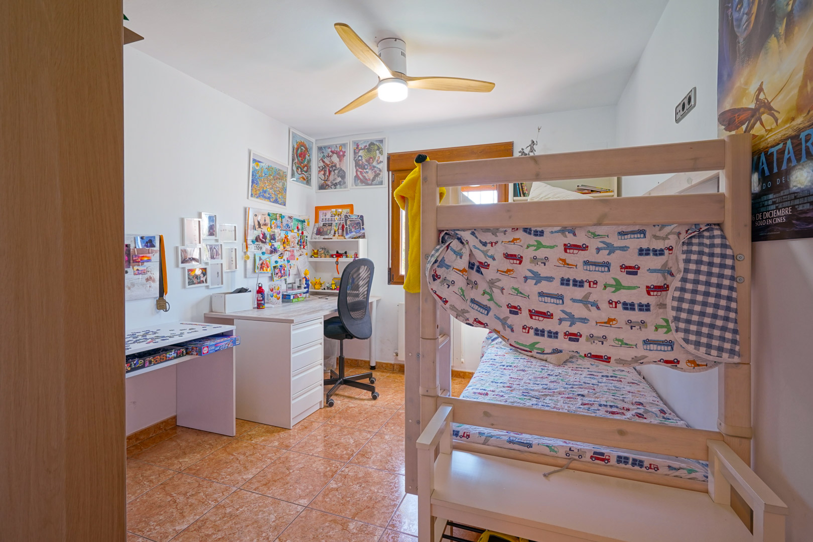 Chambre d'enfant avec lits superposés en bois, un espace bureau et un ventilateur de plafond. La pièce dispose d'un sol en carrelage et d'une belle luminosité naturelle.
