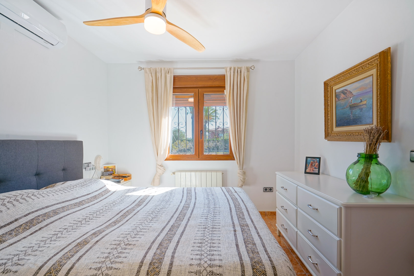 Chambre lumineuse avec fenêtre en bois, ventilateur de plafond à pales en bois, climatisation et commode blanche. Comprend un radiateur et un sol en carrelage.