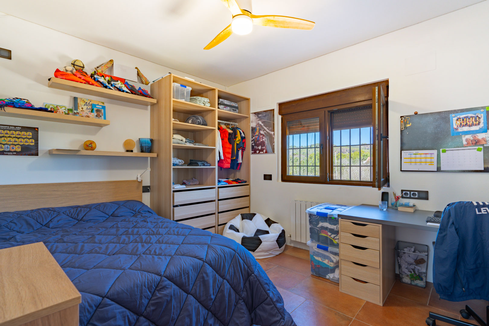 Chambre lumineuse avec sol en terre cuite, espace bureau intégré et rangements ouverts en bois. Une fenêtre à cadre en bois offre une vue dégagée sur le paysage rural.