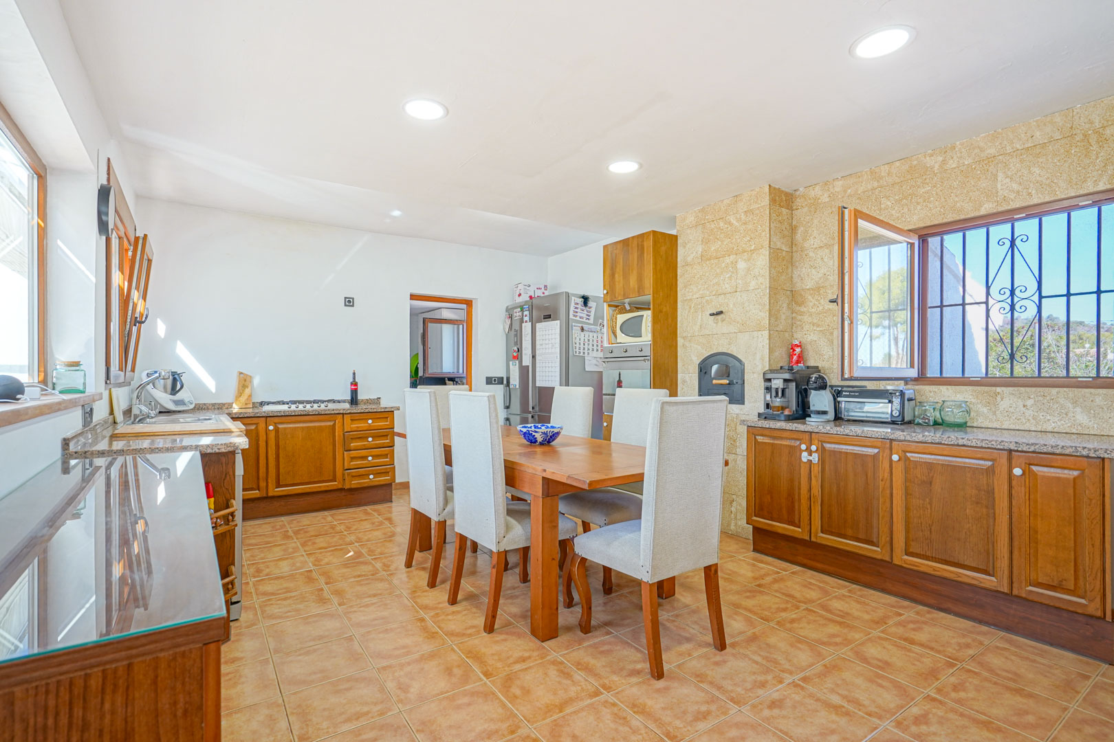 Cuisine rustique avec meubles en bois, plans de travail en granit et four à bois intégré. Comprend un coin repas central avec une grande table et un sol carrelé.