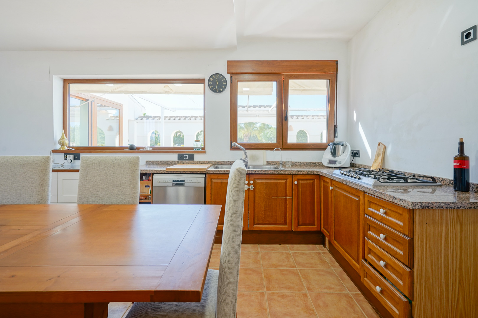 Cuisine rustique avec plans de travail en granit, meubles en bois et coin repas. De grandes fenêtres offrent une vue sur la terrasse extérieure et une lumière naturelle.
