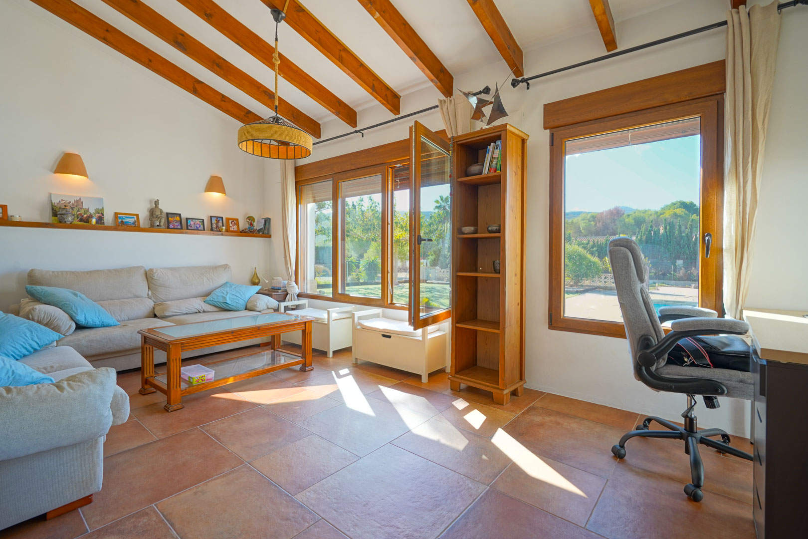 Espace de vie lumineux avec poutres apparentes, sol en terre cuite et coin bureau. Les grandes fenêtres en bois offrent une vue sur le jardin et la piscine.