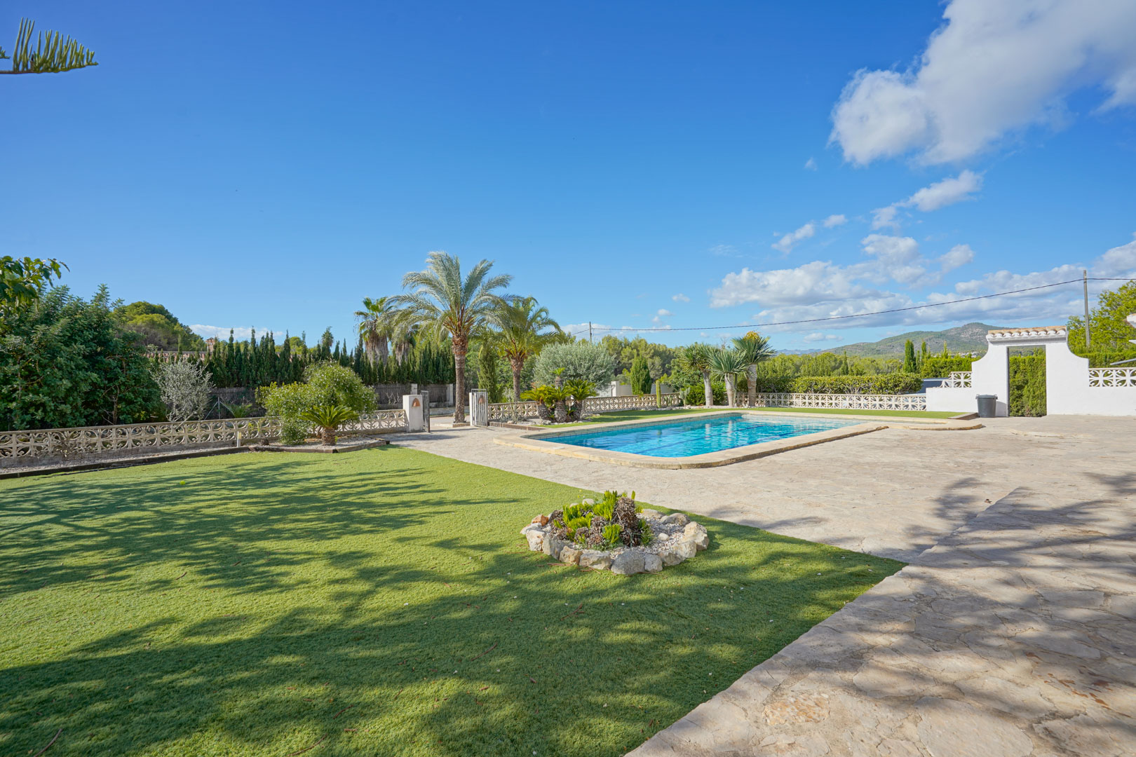 Espace extérieur spacieux avec piscine rectangulaire, terrasse en pierre naturelle et jardin soigné agrémenté de palmiers sous un ciel azur.