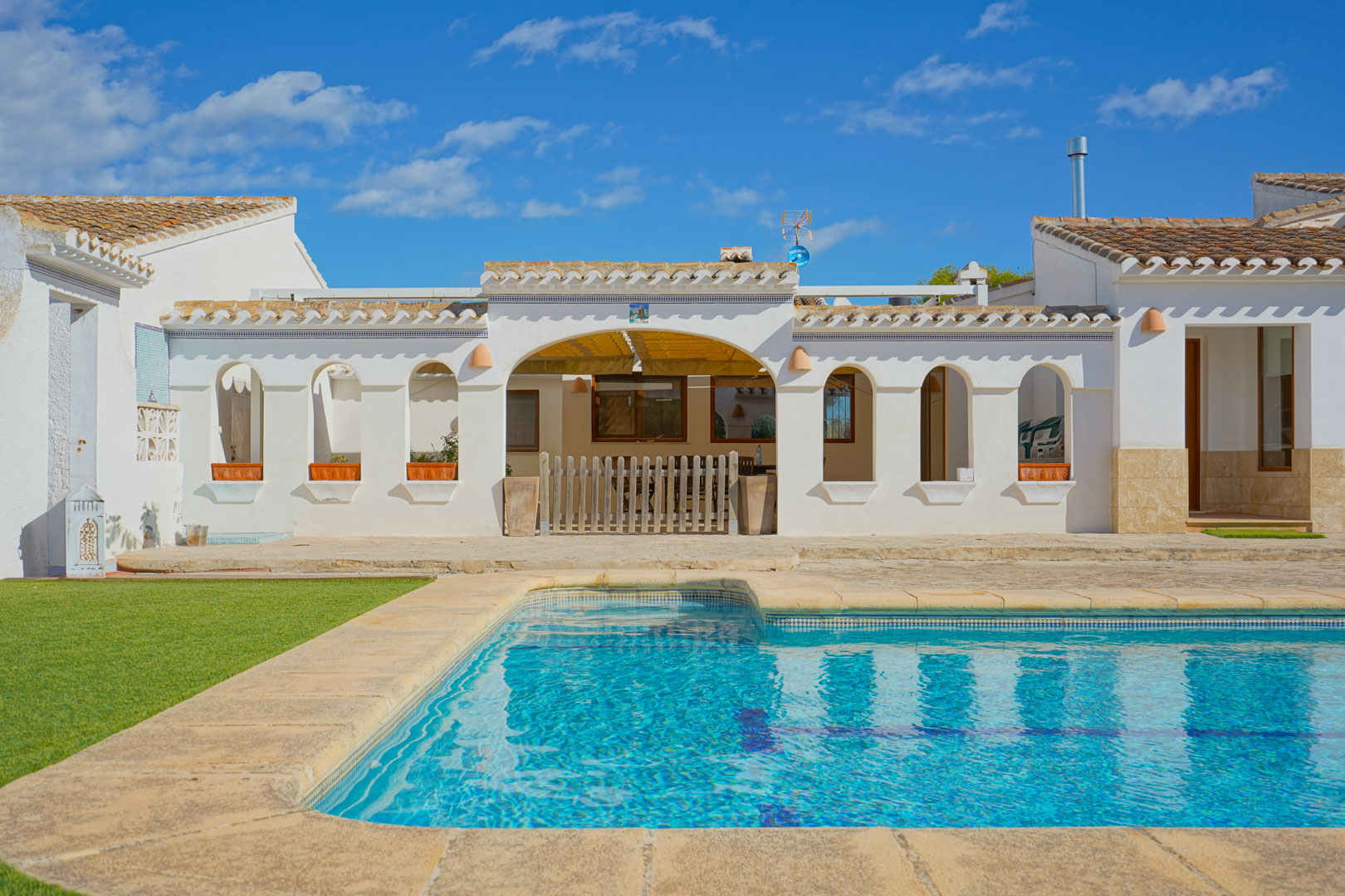 Piscine extérieure avec bordure en pierre naturelle et terrasse ensoleillée face à une façade méditerranéenne blanche aux arches traditionnelles et toit en tuiles.
