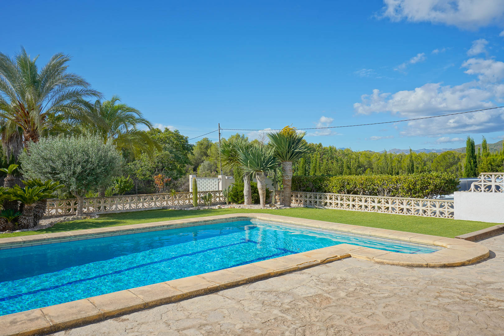Piscine extérieure avec terrasse en pierre naturelle et espace gazonné, entourée de palmiers et d'un mur décoratif en moucharabieh blanc.