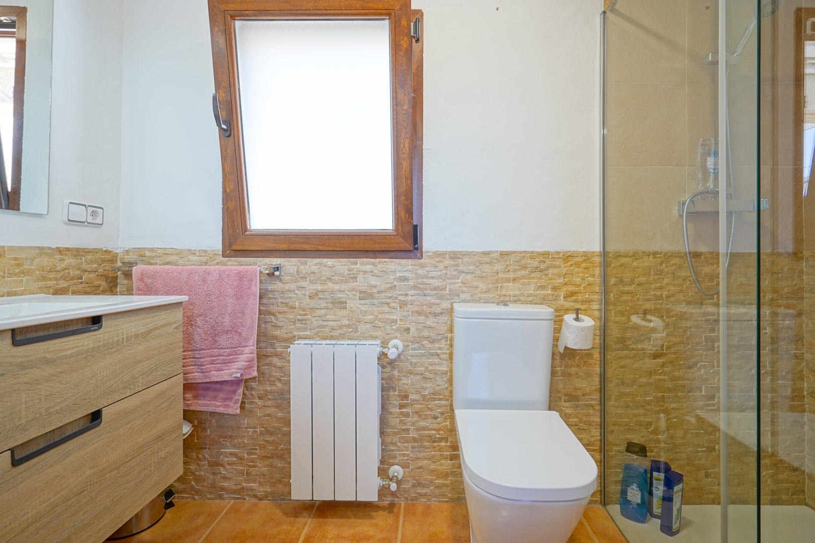 Salle de bain avec meuble vasque en bois, carrelage mural aspect pierre, radiateur blanc et douche à l'italienne avec paroi vitrée sous une fenêtre en bois.