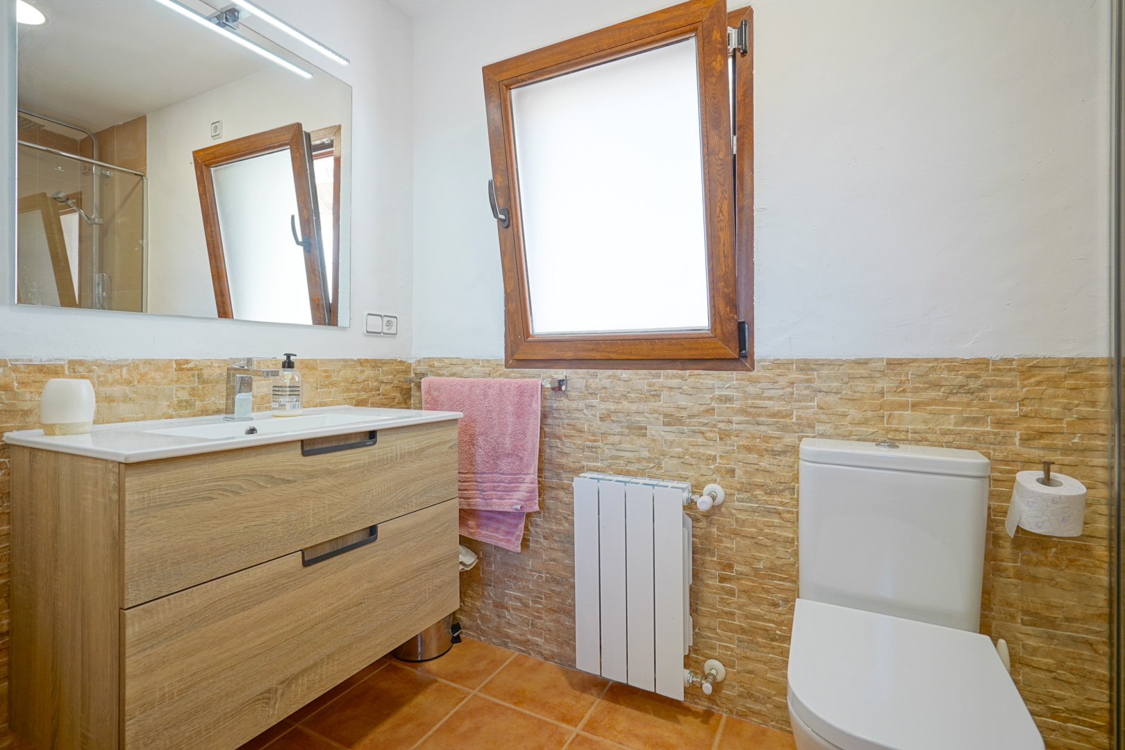 Salle de bain avec meuble vasque en bois, miroir à éclairage LED et carrelage mural effet pierre. Comprend un radiateur et une fenêtre oscillo-battante en bois.
