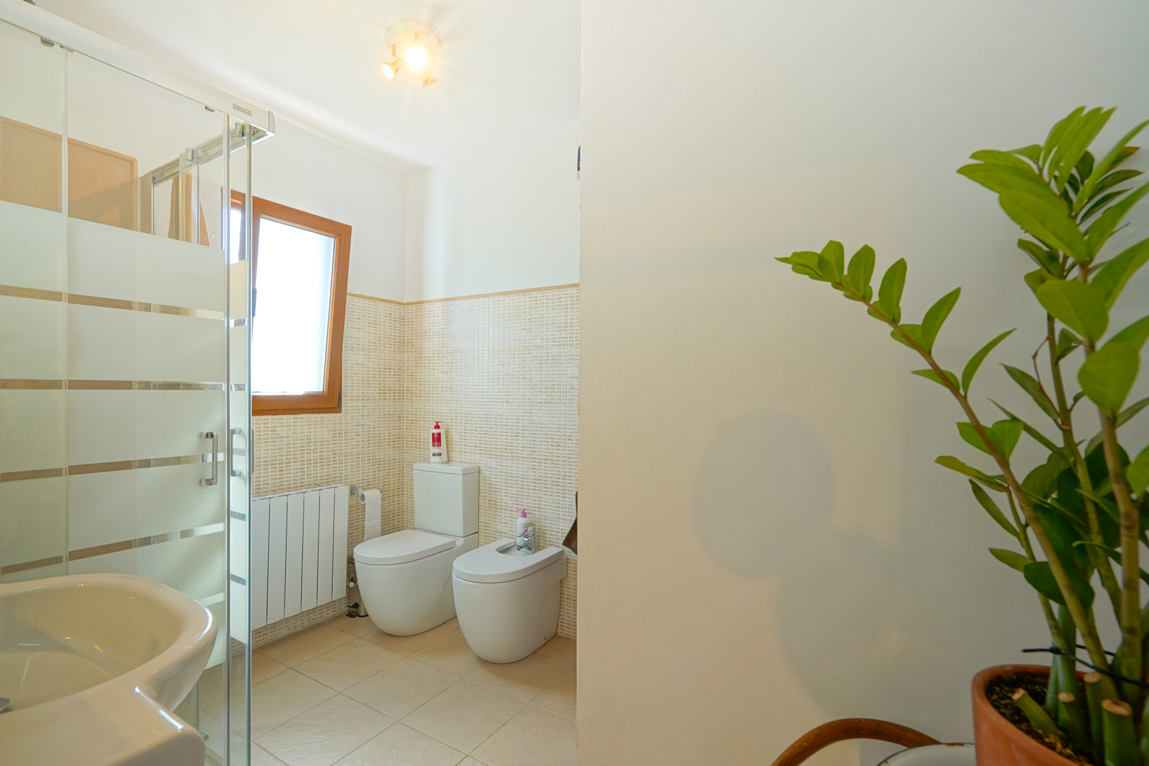 Salle de bains comprenant une douche vitrée, un WC, un bidet et un radiateur, avec des carreaux de mosaïque beige et une fenêtre à cadre en bois.