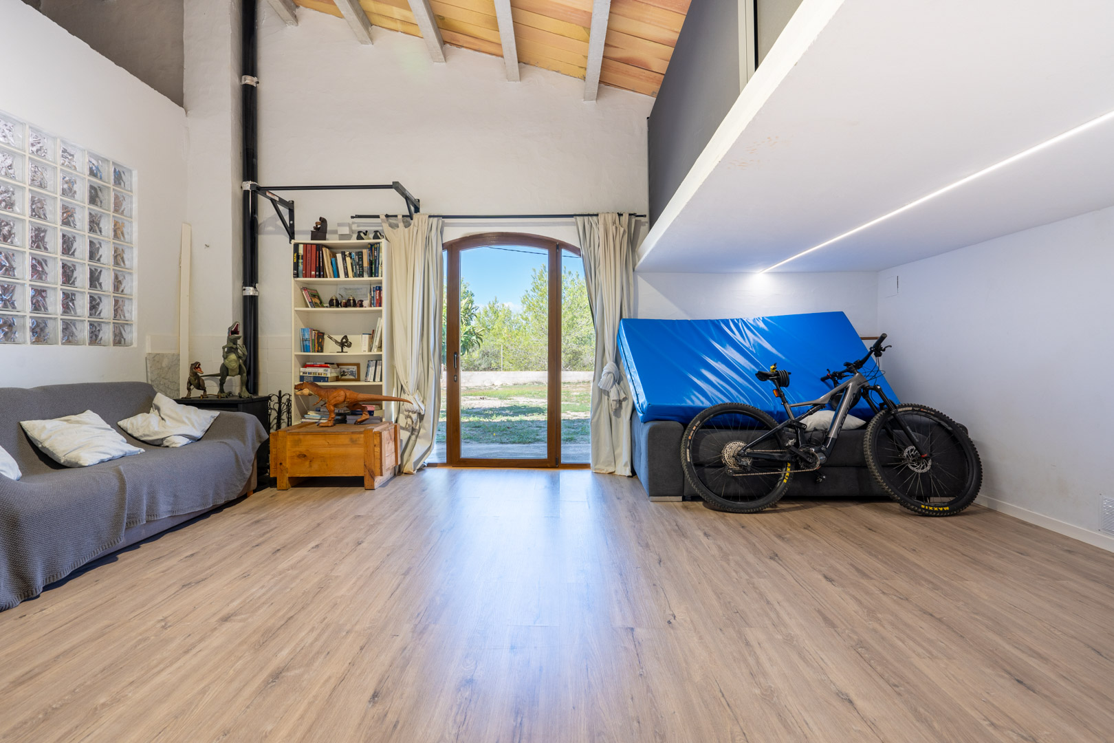 Salle polyvalente avec de hauts plafonds aux poutres apparentes, une mezzanine, un sol stratifié et une porte vitrée cintrée menant à l'extérieur.