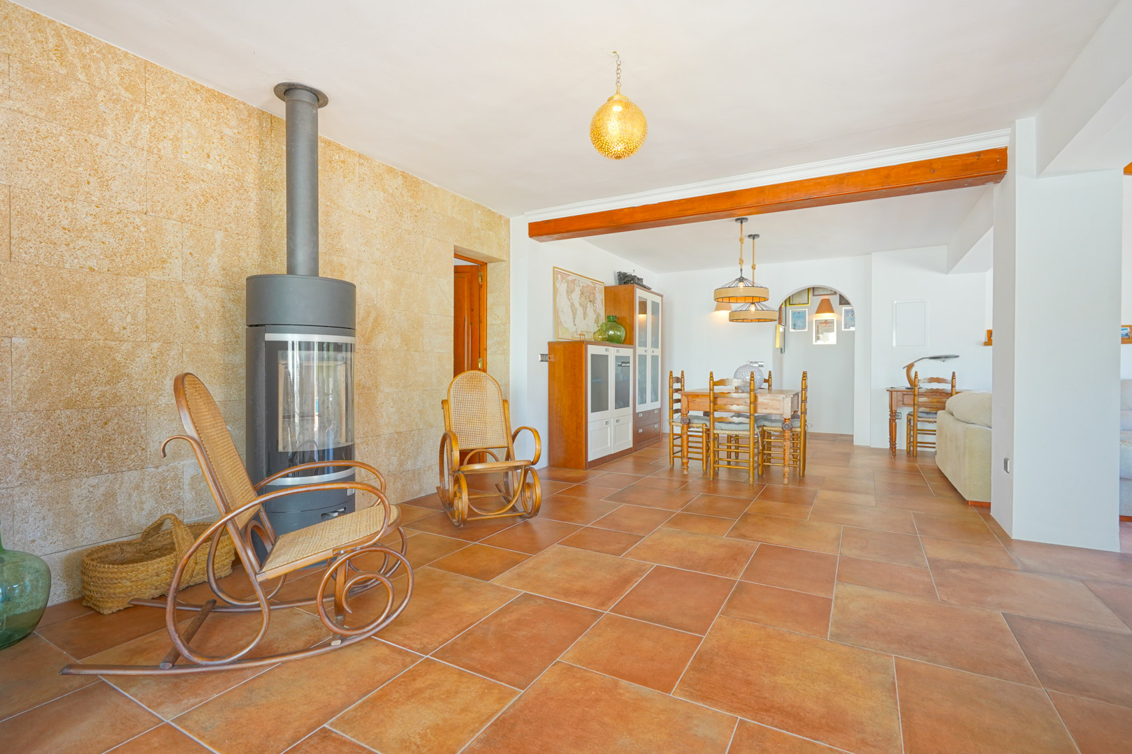 Salon avec poêle à bois moderne sur mur en pierre naturelle, sol en terre cuite et coin repas intégré sous une poutre en bois apparente.