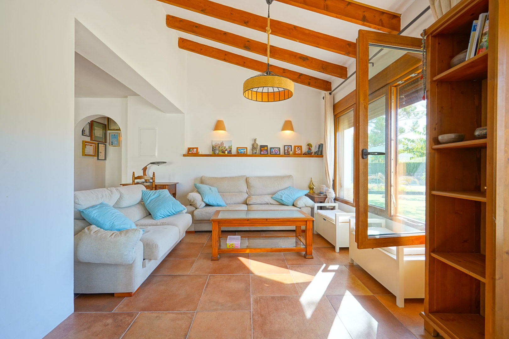 Salon lumineux avec de hauts plafonds à poutres apparentes, un sol en terre cuite et de grandes ouvertures en bois offrant une vue sur le jardin.
