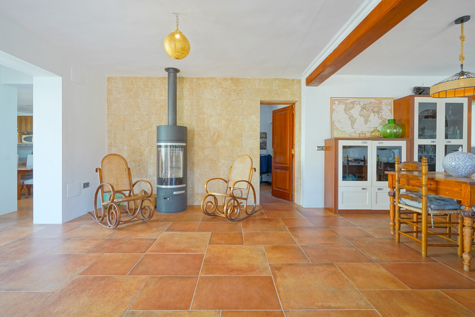 Salon-salle à manger avec sol en terre cuite, poêle à bois moderne sur mur en pierre naturelle et poutres apparentes en bois massif au plafond.