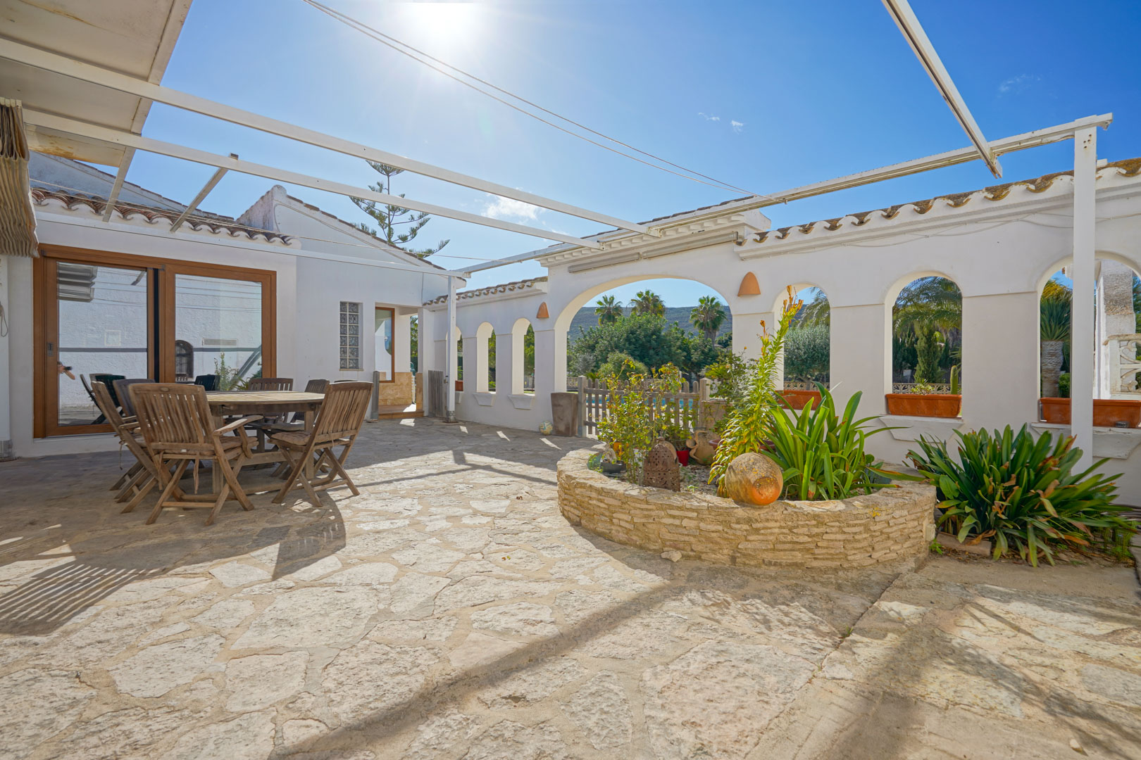 Terrasse extérieure avec sol en pierre naturelle, coin repas en bois et jardinière circulaire sous une structure de pergola métallique.