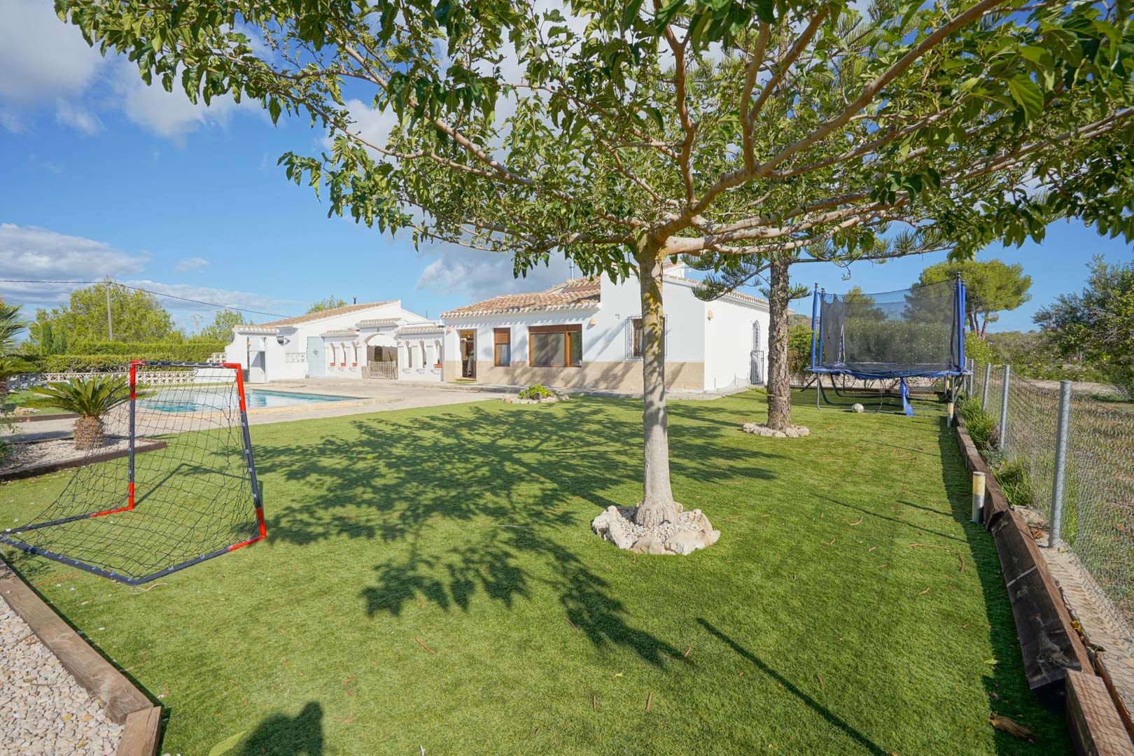 Vaste jardin avec pelouse artificielle et aire de jeux équipée d'un but de foot et d'un trampoline, face à la façade blanche de la villa et la piscine.
