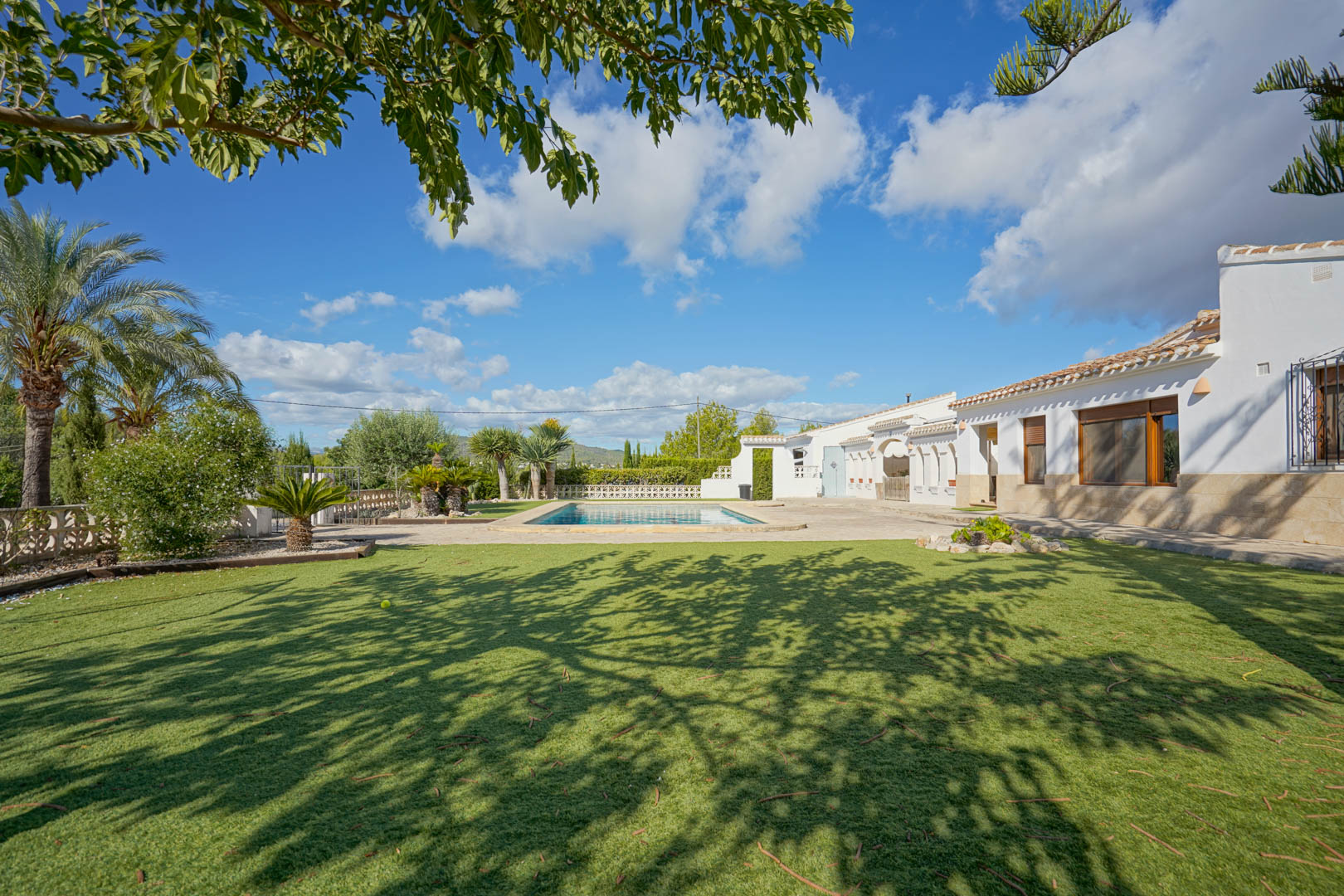 Vue extérieure de la villa aux murs blancs et soubassement en pierre, comprenant une piscine rectangulaire, une terrasse ensoleillée et un jardin avec pelouse.