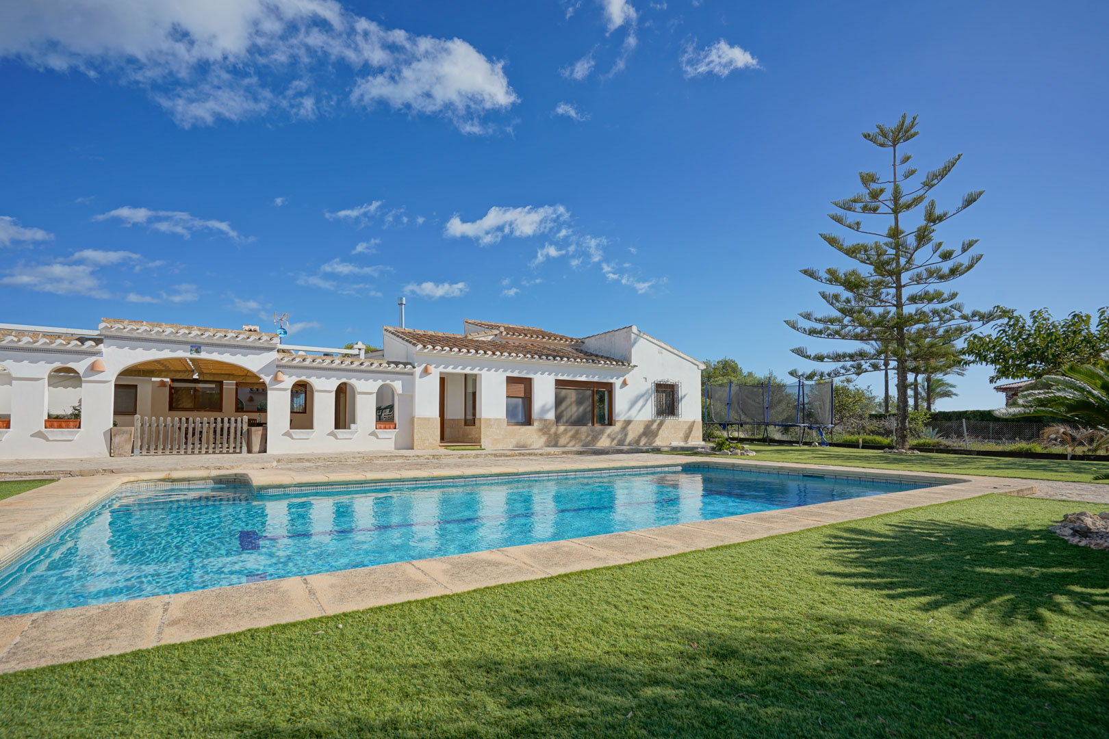 Vue extérieure de la villa avec piscine rectangulaire, pelouse soignée et terrasse en pierre. L'architecture blanche présente des arches traditionnelles et des toits en tuiles.