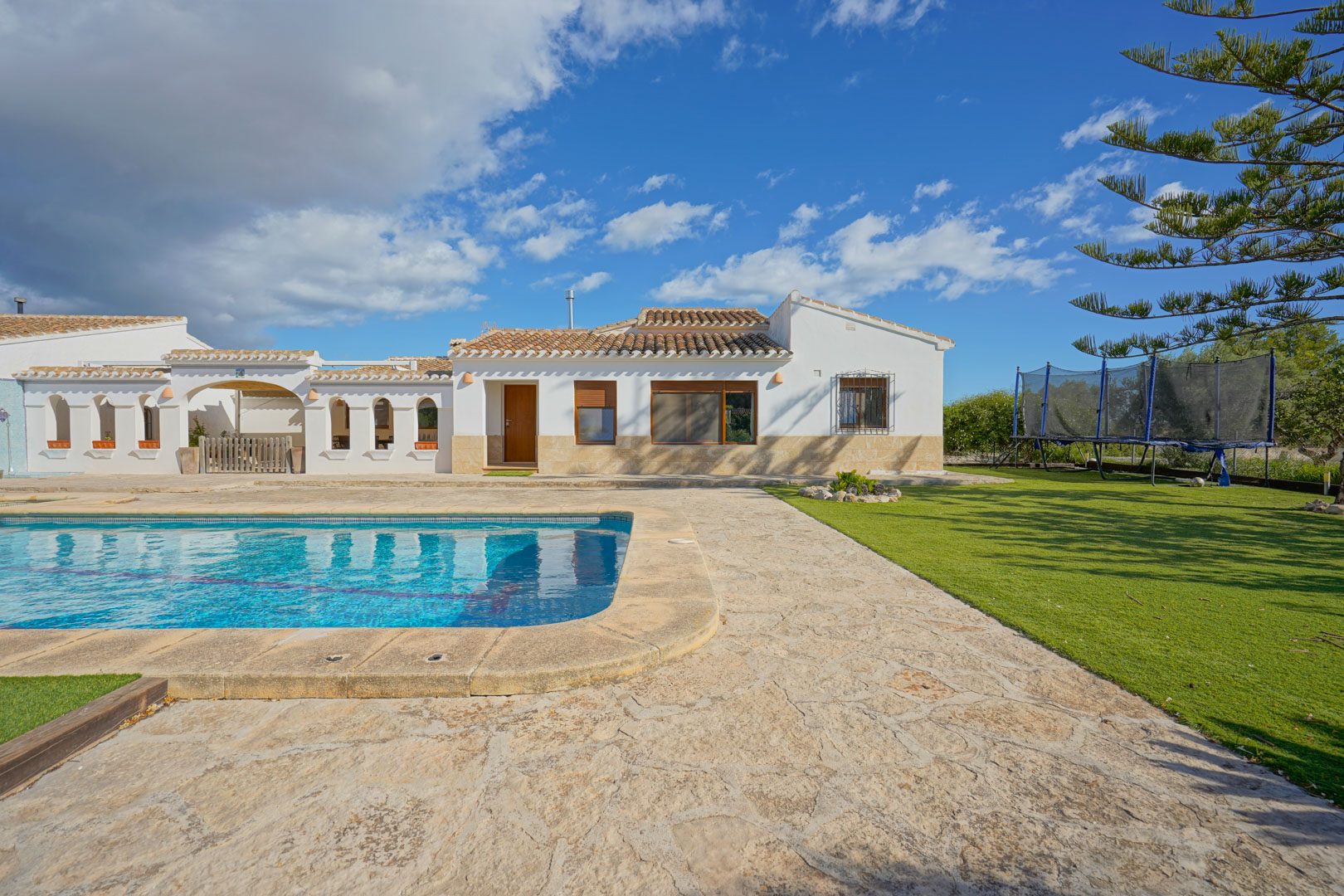 Vue extérieure de la villa avec piscine, terrasse en pierre naturelle et jardin soigné, illustrant l'architecture méditerranéenne sous un ciel dégagé.