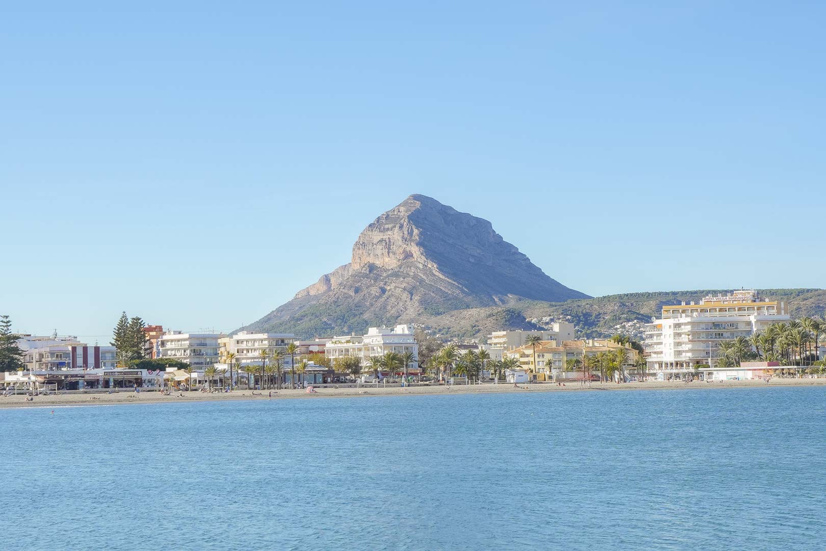 Vue panoramique de la plage de l'Arenal à Jávea, avec le mont Montgó en arrière-plan et les bâtiments côtiers bordant la mer Méditerranée.