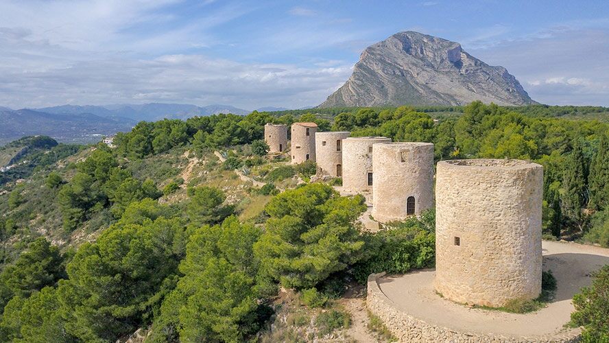 Vue aérienne des moulins en pierre emblématiques de Jávea, avec l'imposant massif du Montgó surplombant le paysage naturel environnant.