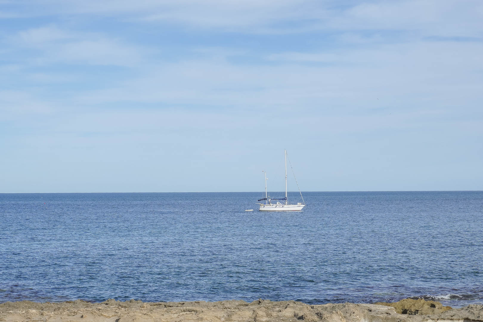 Vues panoramiques sur la mer Méditerranée depuis la côte de Jávea, avec un voilier naviguant près du rivage rocheux sous un ciel dégagé.