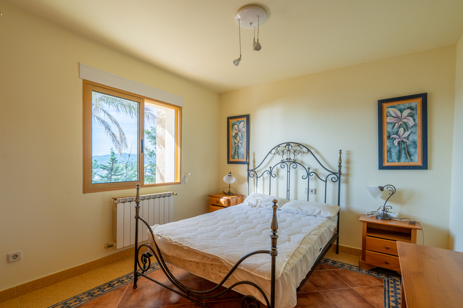 Chambre avec lit en fer forgé, sol en terre cuite avec bordure décorative et fenêtre en bois offrant une vue sur le paysage environnant.