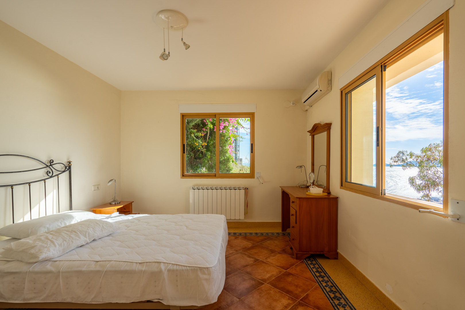 Chambre avec sol en terre cuite et bordures décoratives. Elle dispose de fenêtres en bois avec vue sur mer, d'une tête de lit en fer forgé et de la climatisation.